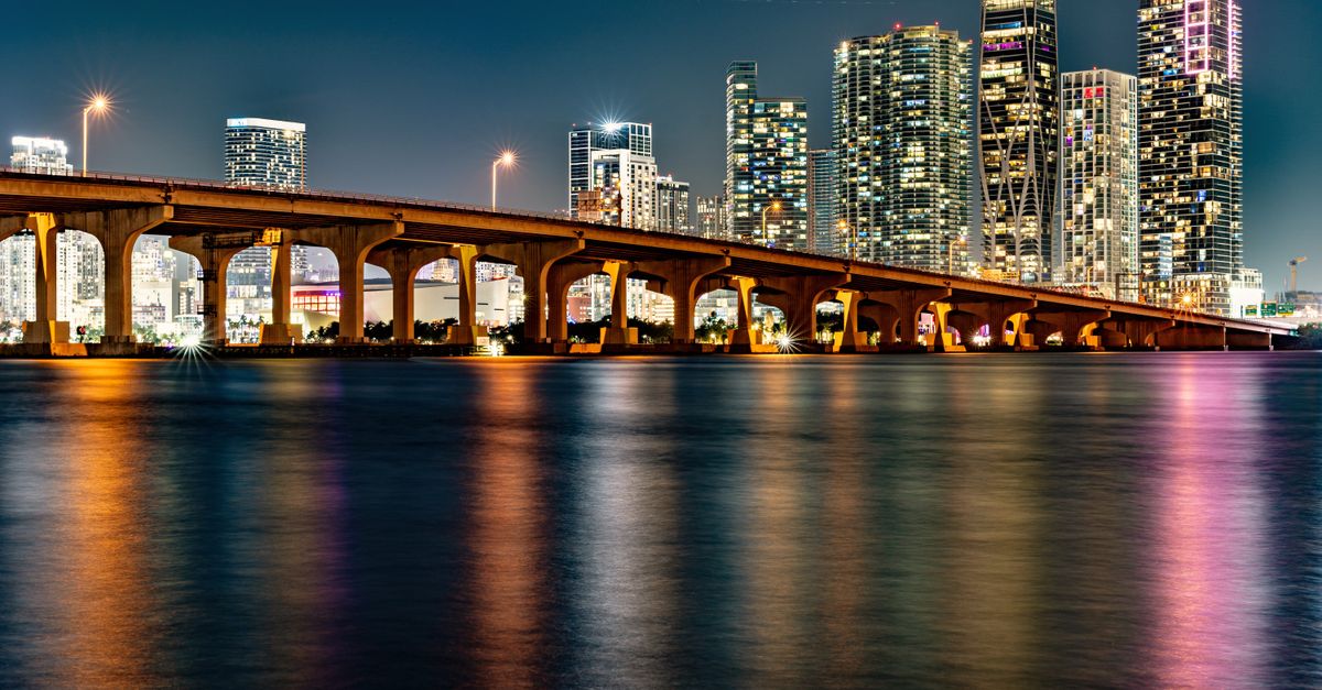 Illuminated Miami skyline with a bridge reflection at night.